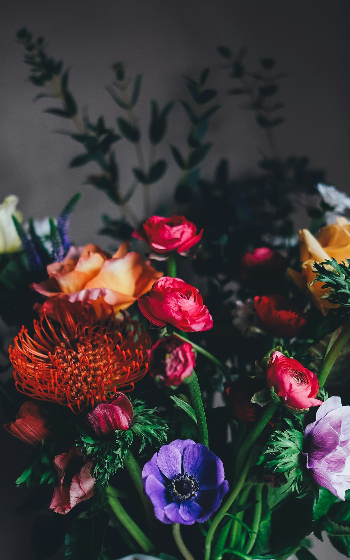 Buchet de atelier cu anemone mov, ranunculus roz, trandafiri somon și leucospermum portocaliu, pe fundal întunecat